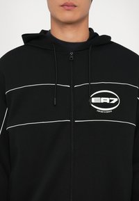 Crna zip-up dukserica s bijelim horizontalnim prugama, s logom na lijevoj strani grudi. Tkanina izgleda glatka s mekim teksturom.