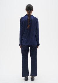 Persona in piedi, voltata di spalle, che indossa una camicia di raso blu navy, jeans blu scuro, scarpe nere e ha lunghi capelli intrecciati che scendono lungo la schiena.