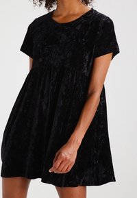 Abito in velluto nero con maniche corte, scollatura arrotondata e vita arricciata. Presenta una texture morbida e una vestibilità ampia e fluida.