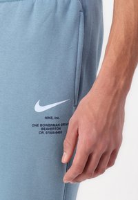 Světle modré kalhoty Nike s hladkým povrchem mají na levém stehně vytištěný bílý znak swoosh a informace o značce.