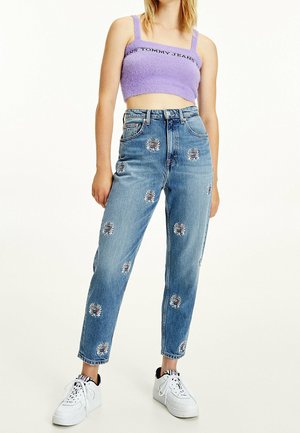 Femme portant un débardeur court violet clair et un jean bleu taille haute avec des patchs circulaires brodés, ainsi que des baskets blanches.