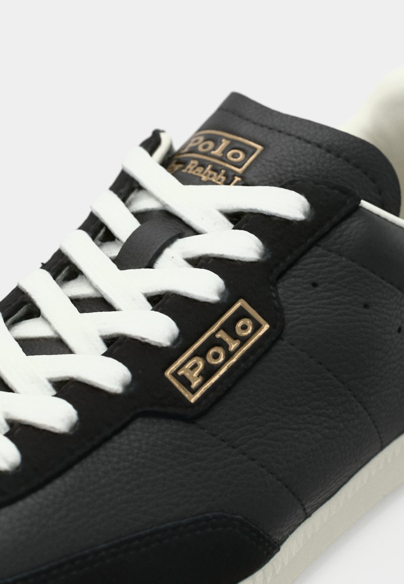 Schwarzer Ledersneaker mit weißen Schnürsenkeln, versehen mit goldenen "Polo"-Logoakzenten und einer strukturierten Oberfläche. Das Design umfasst eine niedrige Silhouette und eine Gummisohle.