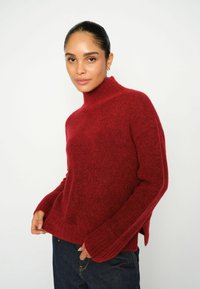 Jersey de punto rojo con cuello alto, mangas largas, aberturas laterales y una textura suave. Lleva puesto sobre unos jeans de mezclilla oscuros, exhibido sobre un fondo blanco.