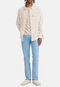 Camicia button-up beige chiaro con due tasche sul petto, t-shirt beige a righe sotto, jeans azzurri chiari e sneakers color crema.