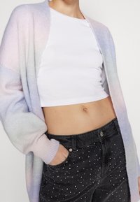 Pastellfärgad gradientkofta i mjukt stickat material, i kombination med en figurnära vit crop top och svarta denimjeans med silverfärgade nitar.