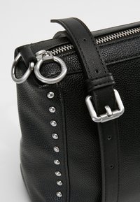 Coin de sac en cuir noir texturé avec fermeture éclair argentée, rivets métalliques, anneau rond et sangle réglable avec boucle argentée.