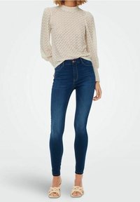 Maglione in crema a maglia texture con collo alto, indossato con jeans skinny blu scuro. Sandali beige chiaro con dettagli di fascette intrecciate.