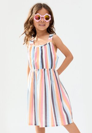 Mädchen trägt ein ärmelloses, mehrfarbiges Kleid mit vertikalen Streifen und rosa, blumenförmige Sonnenbrille, steht mit einer Hand in der Hüfte.
