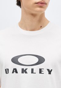 Νεαρό άτομο που φοράει λευκό T-shirt με μεγάλο μαύρο λογότυπο Oakley στο κέντρο του στήθους, στραμμένο ελαφρώς στο πλάι.
