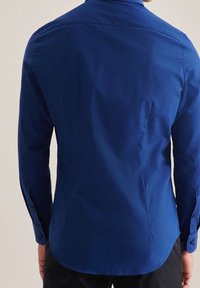 Muž ve fitted, dlouhém rukávu, zapínací košili smaragdově modré barvy, zobrazený zezadu na neutrálním pozadí.