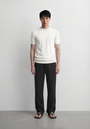 Νέος άνδρας που στέκεται μπροστά σε έναν λευκό τοίχο, φοράει λευκό κοντομάνικο πουκάμισο, μαύρο παντελόνι και μαύρα σαγιονάρες, κοιτάζοντας μπροστά με ουδέτερη έκφραση.