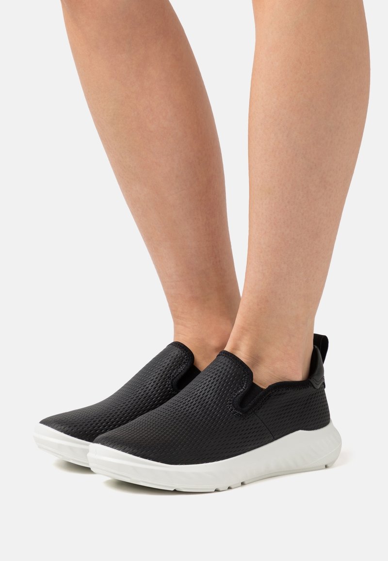 Jambes portant des baskets noires à enfiler avec des dessus texturés et des semelles en caoutchouc blanches sur un fond clair.