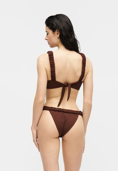 Femme aux longs cheveux noirs vue de dos, portant un bikini marron foncé texturé avec des bretelles froncées et une fermeture nouée dans le dos.