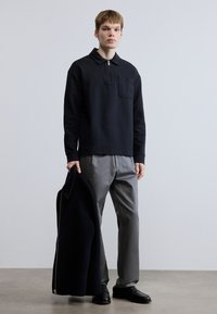 Μαύρο μακρυμάνικο πόλο μπλουζάκι με φερμουάρ στο γιακά και τσέπη, συνδυασμένο με γκρι παντελόνι. Το μοντέλο κρατάει ένα σκούρο σακάκι, στέκεται σε ουδέτερο φόντο.