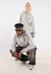 Helle graue Hoodies mit frontaler bestickter, farbenfroher Gestaltung, getragen von zwei Models. Das männliche Model trägt außerdem schwarze Sonnenbrillen und ausgewaschene Jeans.