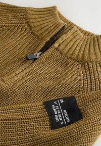 Gestrickter Pullover in Senfgelb, mit einem gerippten Kragen, halbem Reißverschluss und einem Stoffetikett mit Pflegehinweisen. Texturierte Details sind sichtbar.