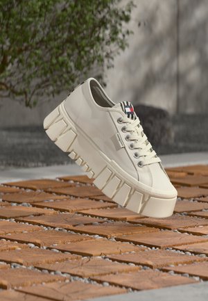 Béžová teniska s masivní podrážkou a výrazným vzorkem, kovovými očky, značkovou jazykovou štítkem a logem Tommy Hilfiger, která visí nad dřevěnými dlaždicemi venku.