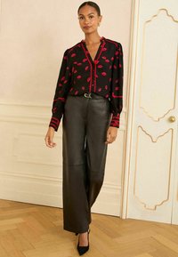 Blusa nera con stampe di labbra rosse, colletto arricciato e chiusura con bottoni; abbinata a pantaloni in pelle nera a vita alta e tacchi.
