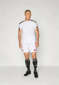 Hvit idrettsgenser med svarte striper på skuldrene, kombinert med hvite shorts med røde aksenter og en logo, svarte leggbeskyttelser og fotballsko.