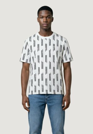 Uomo con indosso una camicia bianca a maniche corte con motivi a righe verticali nere e jeans blu, in piedi contro uno sfondo semplice.