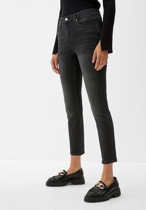 Persoon met strakke zwarte jeans, kort boven de enkels, gecombineerd met zwarte chunky loafers met een grote gouden kettingdetail.