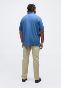 Άνδρας όρθιος με την πλάτη facing φοράει μπλε πόλο με κοντά μανίκια, μπεζ παντελόνι και μαύρα παπούτσια σε απλό ανοιχτό φόντο.