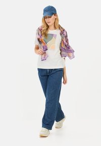Hvid t-shirt med abstrakt grafik, parret med blå denimjeans, en blå kasket og et flerfarvet gennemsigtigt tørklæde draperet omkring skuldrene.