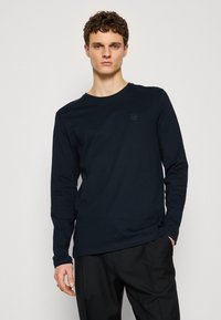 Camiseta de manga larga azul marino en algodón suave con escote redondo y un pequeño parche de logo en forma de cuadrado en el pecho, combinada con pantalones negros.