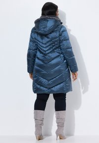 Cappotto invernale blu, imbottito, con motivo a zigzag, cappuccio foderato in pelliccia e maniche lunghe, abbinato a stivali grigi fino al ginocchio e pantaloni scuri.