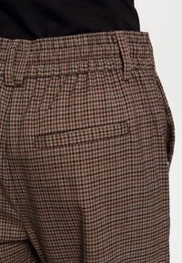 Bruna rutiga byxor i en texturerad tyg, med en bakficka och en strukturerad midja. Mönstret består av små rutor i mörkare nyanser.