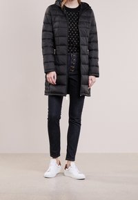 Czarna kurtka puchowa z kapturem, o pikowanej konstrukcji, z zapięciem na zamek i kieszeniami po bokach. Noszona na czarnym swetrze i ciemnych dżinsach.