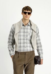 Koszula w kratę z kołnierzykiem zapinanym na guziki, beżowy sweter dziany narzucony na ramiona, brązowe spodnie sztruksowe oraz brązowa skórzana torba trzymana w ręce.