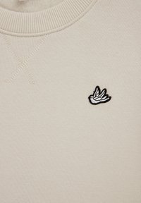 Bluză sport bej deschis din amestec de bumbac, cu guler rotund texturat și un mic logo brodat alb-negru cu o pasăre pe piept.