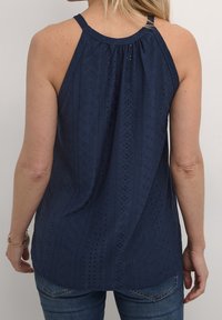 Kvinna iförd ärmlös marinblå ögonstickad topp och blå jeans, sedd bakifrån mot en enkel vit bakgrund.