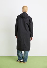 Svart lang regnfrakk med hette, laget av vanntett materiale, med en logo på ryggen og knappløsninger ved ermene, kombinert med blå jeans og sneakers.