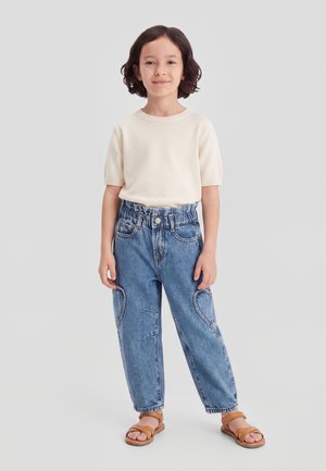 Kind mit kurzen dunklen Haaren trägt ein cremefarbenes kurzärmeliges Oberteil, hoch taillierte blaue Jeans mit Herz-Patches und beige Sandalen, steht vor einem grauen Hintergrund.