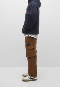 Person iført navy hoodie, hvid t-shirt, brune cargo bukser med klaplommer og hvide sneakers med sorte detaljer, der står sidelæns.