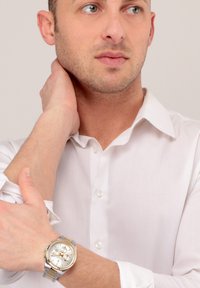 Silberne und goldene Armbanduhr mit rundem Ziffernblatt, drei kleinen Zifferblättern, einem strukturierten Gehäuse und einem Metall-Gliederarmband. Getragen über einem weißen Hemd mit Knopfleiste.