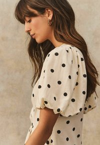 Cremfarbene Bluse mit kurzen, gepufften Ärmeln und schwarzen Punkten. Weicher Stoff mit glatter Textur; minimalistisches Design mit rundem Ausschnitt.