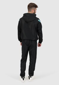 Zwarte sportieve trainingspak met capuchon, voorzien van lichtblauwe schouderaccenten en een gladde textuur. Schoenen zijn wit met zwarte details.