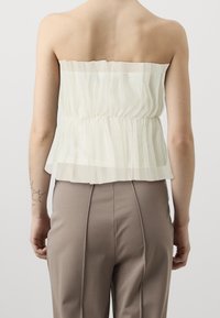 Top senza spalline in tessuto bianco trasparente con vita arricciata, caratterizzato da un design a strati testurizzato. Abbinato a pantaloni beige sagomati.