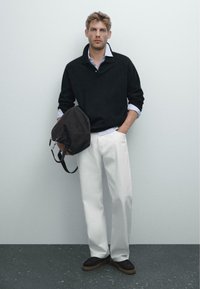 Schwarzer Strickpullover mit Kragen, weißes Hemd mit Kragen, weiße weit geschnittene Hose, dunkelbraune Tasche und schwarze Schuhe mit hellbraunen Sohlen.
