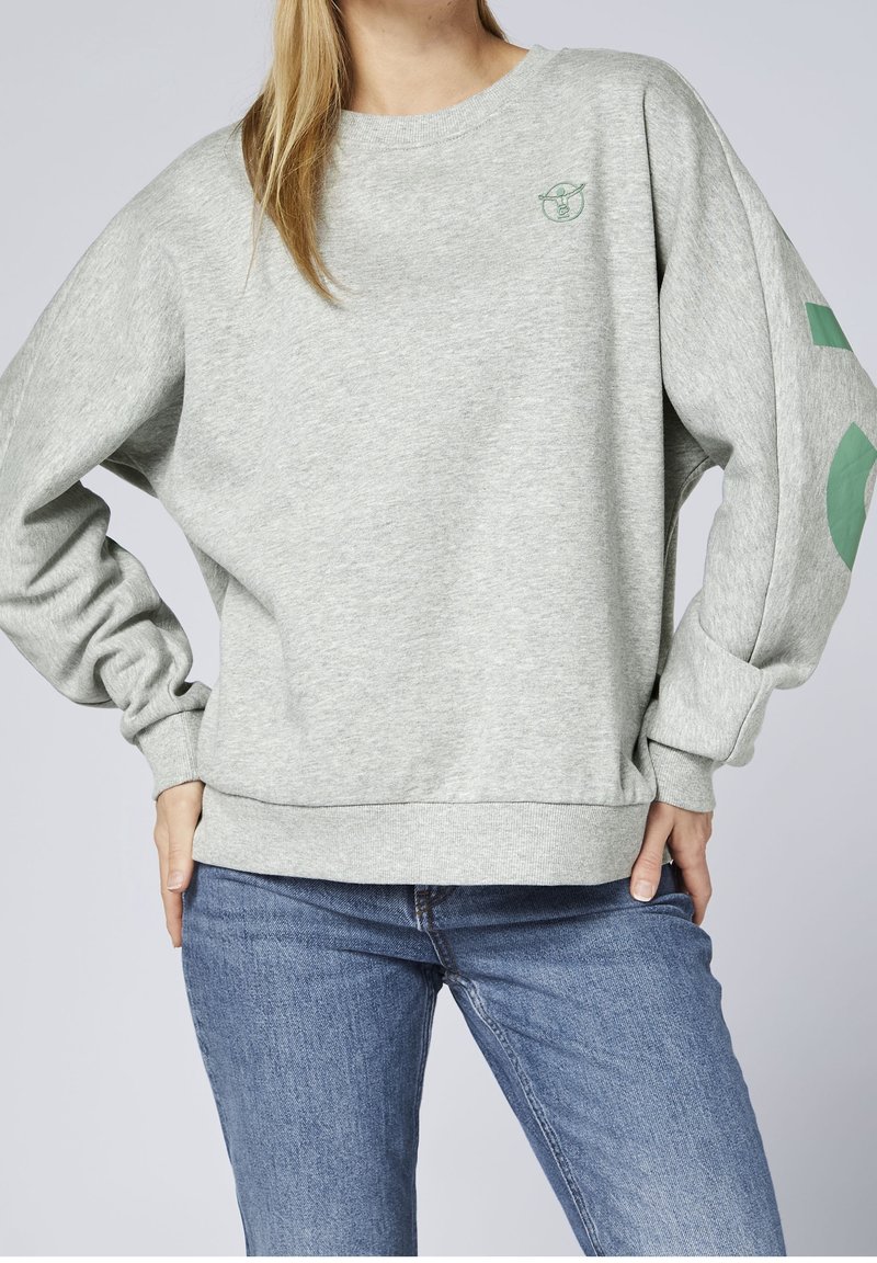 Personne portant un sweat-shirt gris clair avec un petit logo vert sur la poitrine et un design géométrique vert sur la manche, associé à un jean bleu.