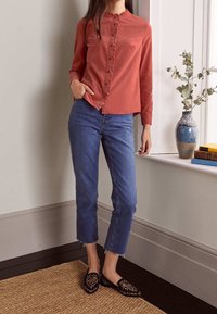 Camicia button-up color ruggine con dettagli di volant, abbinata a jeans blu alla caviglia e scarpe slip-on nere con un motivo distintivo.
