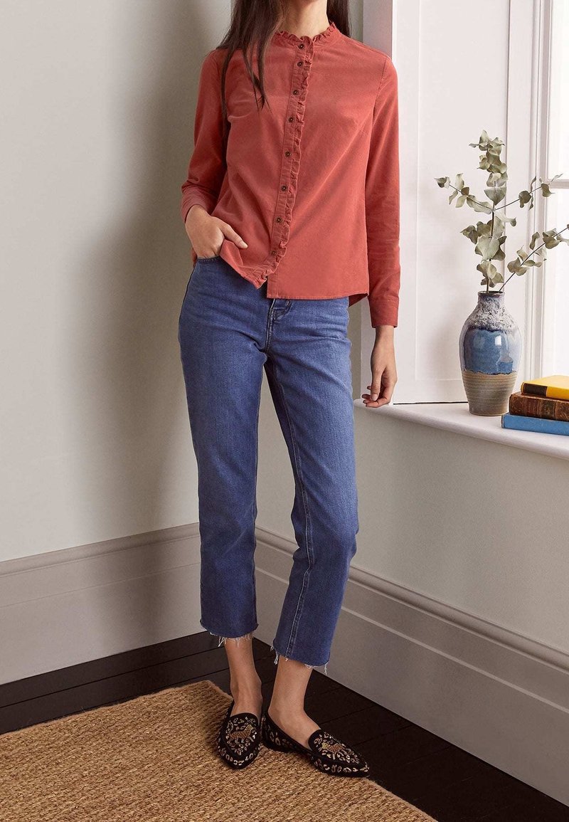 Camicia button-up color ruggine con dettagli di volant, abbinata a jeans blu alla caviglia e scarpe slip-on nere con un motivo distintivo.