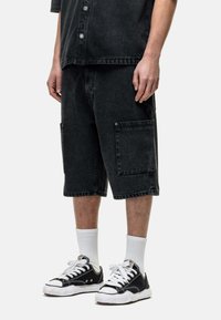Shorts cargo en denim noir avec poches latérales, longueur genou, conçus avec une coupe décontractée. Associés à des baskets noires et des chaussettes blanches.