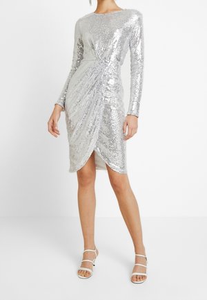 Kobieta ubrana w srebrną, dopasowaną sukienkę z długim rękawem, zdobioną cekinami, z marszczonym tulipanowym dołem oraz białe, odkryte buty na obcasie z paskami, stojąca na białym tle.