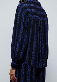 Schwarzes Hemd mit floralem Muster und blauen Akzenten, mit langen Ärmeln, plissiertem Rücken und klassischem Kragen. Weiche Stoffstruktur.