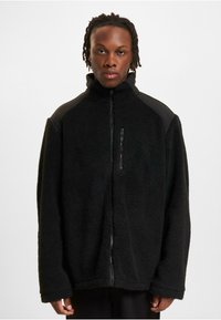 Schwarze Fleecejacke mit hohem Kragen, Reißverschluss vorne, strukturierter Stoff und schwarzen Schulterakzenten. Lange Ärmel mit lockerem Sitz.