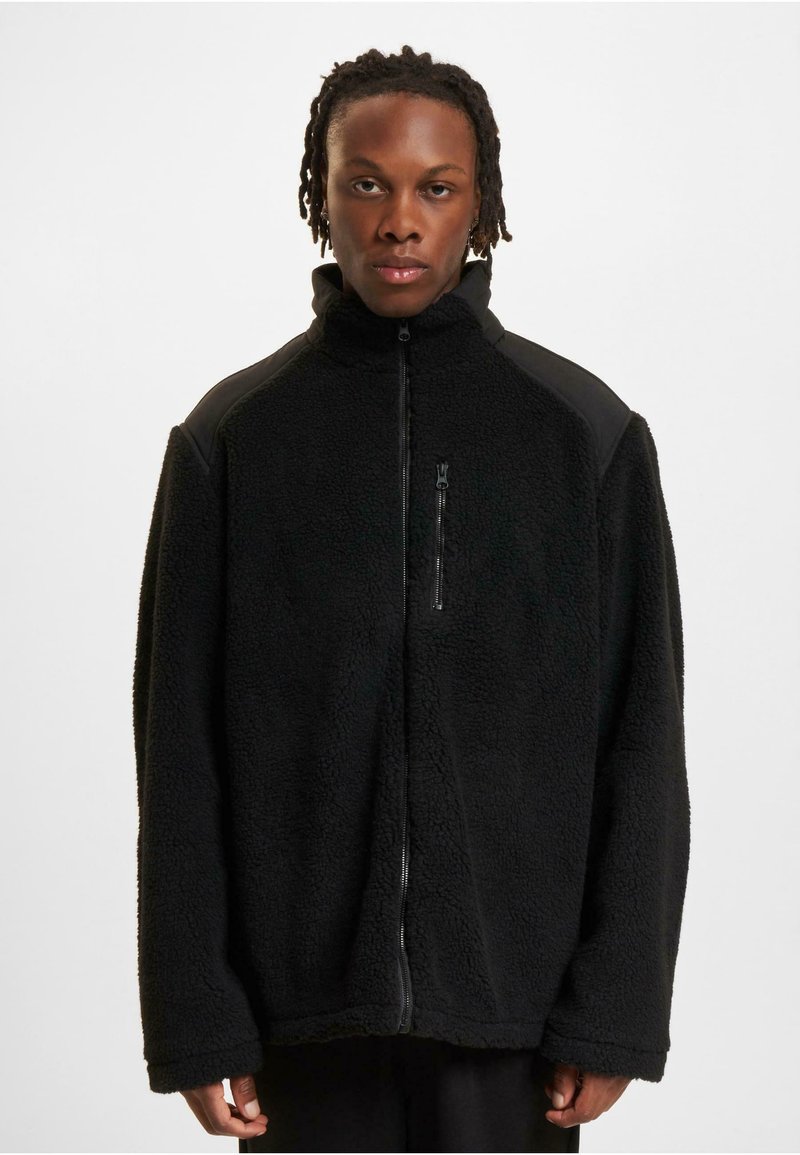 Schwarze Fleecejacke mit hohem Kragen, Reißverschluss vorne, strukturierter Stoff und schwarzen Schulterakzenten. Lange Ärmel mit lockerem Sitz.